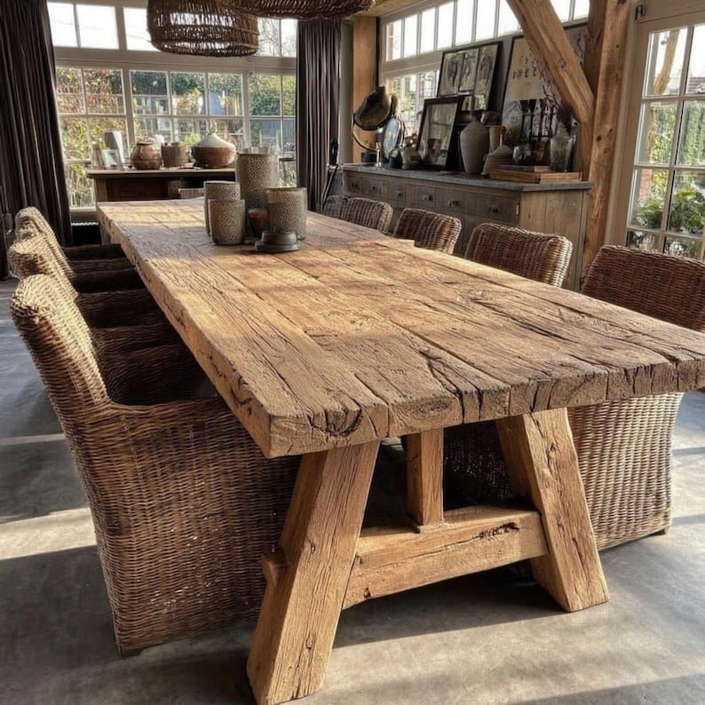 May include: A large, rustic wooden dining table with a natural finish. The table has a thick, rectangular top and sturdy A-frame legs. Woven wicker chairs surround the table. The room has large windows and decorative items.