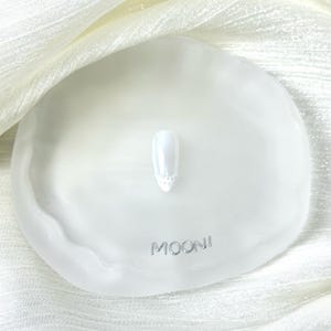 Puede incluir: Una u&ntilde;a blanca con un acabado nacarado y un dise&ntilde;o decorativo con joyas en la base. La u&ntilde;a se muestra en una bandeja blanca de forma org&aacute;nica con la palabra "MOONI" en letras plateadas. El fondo es una tela blanca y sedosa.
