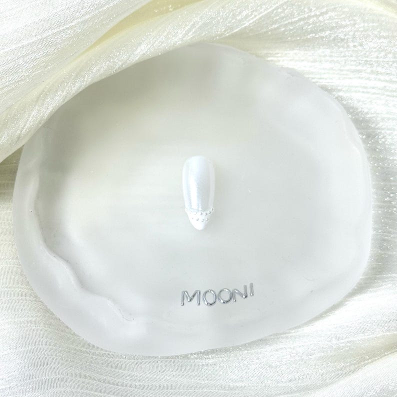 Puede incluir: Una u&ntilde;a blanca con un acabado nacarado y un dise&ntilde;o decorativo con joyas en la base. La u&ntilde;a se muestra en una bandeja blanca de forma org&aacute;nica con la palabra "MOONI" en letras plateadas. El fondo es una tela blanca y sedosa.
