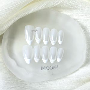 Puede incluir: Un conjunto de u&ntilde;as postizas blancas con dise&ntilde;o de manicura francesa. Cada u&ntilde;a tiene un acabado nacarado y est&aacute; adornada con un peque&ntilde;o adorno decorativo cerca de la cut&iacute;cula. Las u&ntilde;as est&aacute;n dispuestas sobre una base transparente de forma ovalada con la marca "MOONI".