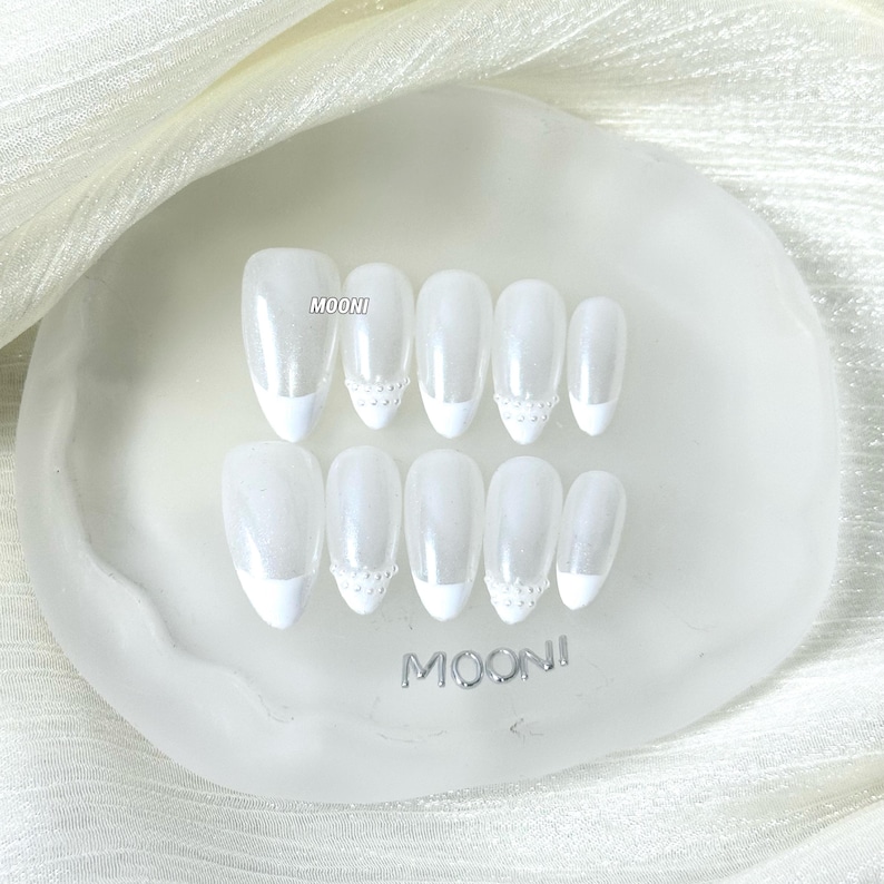 Puede incluir: Un conjunto de u&ntilde;as postizas blancas con dise&ntilde;o de manicura francesa. Cada u&ntilde;a tiene un acabado nacarado y est&aacute; adornada con un peque&ntilde;o adorno decorativo cerca de la cut&iacute;cula. Las u&ntilde;as est&aacute;n dispuestas sobre una base transparente de forma ovalada con la marca "MOONI".