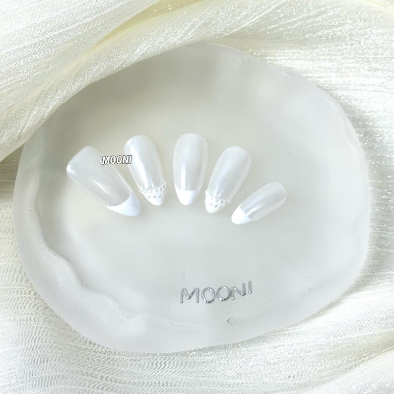 Puede incluir: Cinco u&ntilde;as artificiales blancas nacaradas con puntas blancas y detalles decorativos se muestran sobre una superficie blanca texturizada. La palabra "MOONI" es visible en la superficie. Las u&ntilde;as est&aacute;n dispuestas sobre una superficie blanca texturizada.
