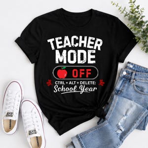 May include: Black t-shirt with the text "TEACHER MODE OFF" in white, with a red apple graphic. Below the apple is "CTRL + ALT + DELETE: School Year". White sneakers and blue jeans are visible.