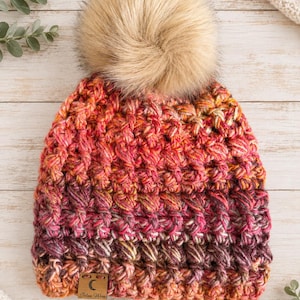 May include: A chunky knit beanie featuring a variegated yarn pattern in shades of orange, pink, and deep purple. The hat is topped with a large, fluffy beige faux fur pom-pom and includes a small rectangular wooden tag with a crescent moon logo.