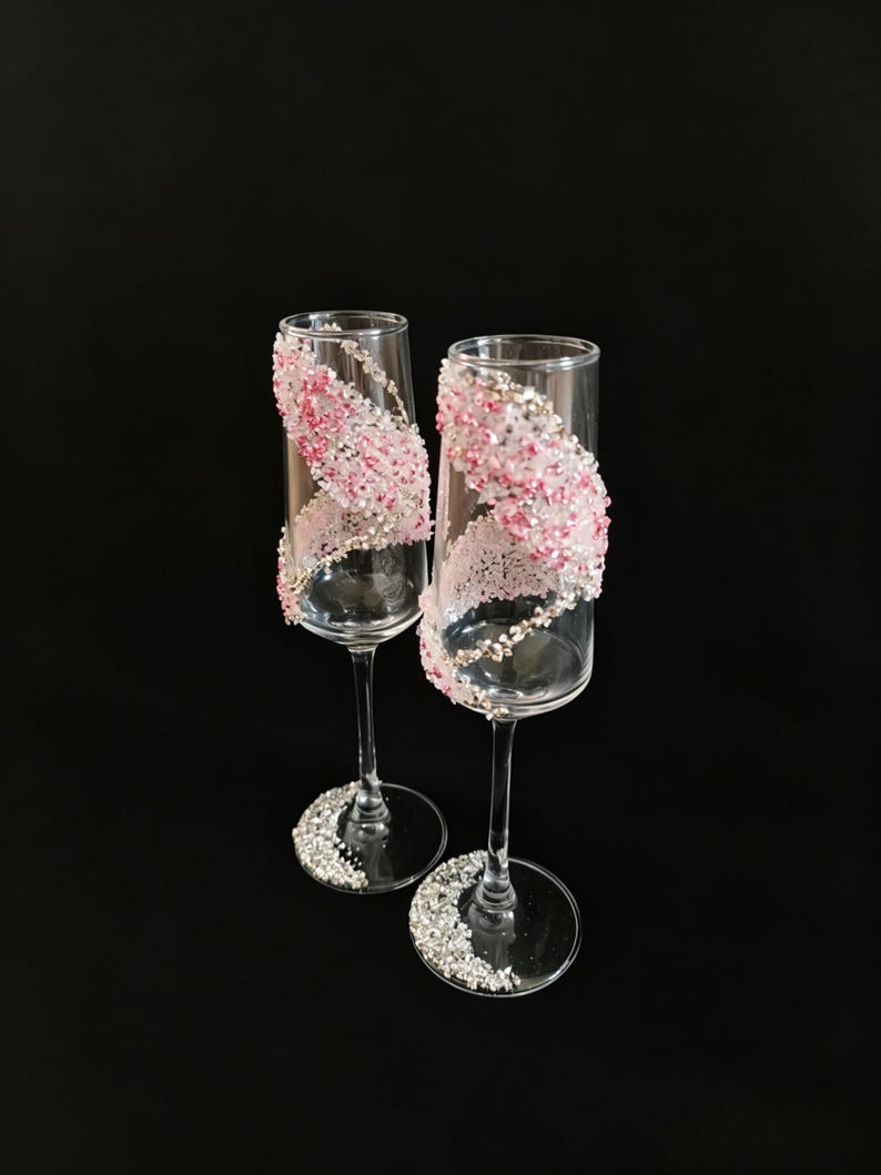 K&ouml;nnte beinhalten: Zwei Sektgl&auml;ser mit klarem Glas und einem dekorativen Design. Das Design zeigt rosa und silberne Verzierungen, die sich um das Glas winden. Der Fu&szlig; jedes Glases ist ebenfalls mit silbernen Verzierungen verziert.