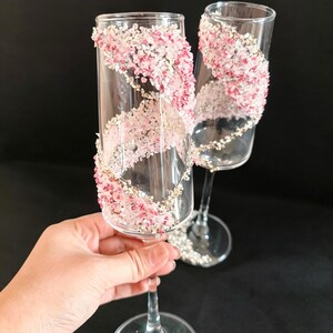 K&ouml;nnte beinhalten: Zwei klare Sektgl&auml;ser, verziert mit rosa und wei&szlig;en floralen Akzenten, die sich &uuml;ber das Glas spiralf&ouml;rmig erstrecken. Der Fu&szlig; jedes Glases ist mit silbernen Glitzer verziert. Ideal f&uuml;r eine Hochzeit oder einen besonderen Anlass.