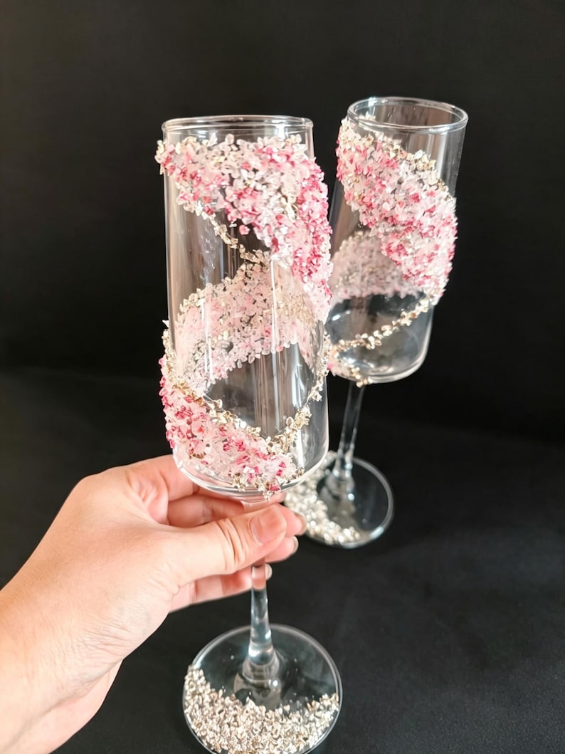 K&ouml;nnte beinhalten: Zwei klare Sektgl&auml;ser, verziert mit rosa und wei&szlig;en floralen Akzenten, die sich &uuml;ber das Glas spiralf&ouml;rmig erstrecken. Der Fu&szlig; jedes Glases ist mit silbernen Glitzer verziert. Ideal f&uuml;r eine Hochzeit oder einen besonderen Anlass.