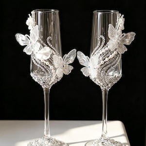 May include: Two clear champagne flutes decorated with white butterflies, pearls, and rhinestones. The butterfly and pearl design adorns the glass, and the base is embellished with rhinestones. The flutes are set against a black background.