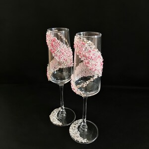 K&ouml;nnte beinhalten: Zwei Sektgl&auml;ser mit klaren Glasschalen und Stielen. Jedes Glas ist mit einem wirbelnden Muster aus rosa und silbernen Verzierungen verziert. Der Fu&szlig; jedes Glases ist ebenfalls mit silbernen Verzierungen bedeckt. Der Hintergrund ist schwarz.