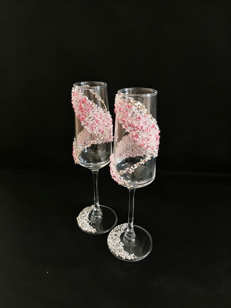 K&ouml;nnte beinhalten: Zwei Sektgl&auml;ser mit klaren Glasschalen und Stielen. Jedes Glas ist mit einem wirbelnden Muster aus rosa und silbernen Verzierungen verziert. Der Fu&szlig; jedes Glases ist ebenfalls mit silbernen Verzierungen bedeckt. Der Hintergrund ist schwarz.