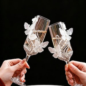 May include: Two champagne flutes clink together, filled with a golden liquid. Each glass is adorned with white butterflies, pearls, and sparkling rhinestones. The stem and base are also decorated with rhinestones and pearls, set against a black background.