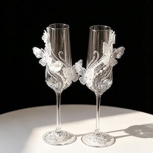 May include: Two clear champagne flutes with intricate embellishments. Each glass features white butterfly and floral accents, along with sparkling rhinestones and pearls. The stems and bases are also adorned with rhinestones, creating an elegant look.