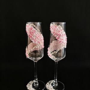 K&ouml;nnte beinhalten: Zwei Sektgl&auml;ser mit klarem Glas und einem dekorativen Design. Das Design zeigt eine Spirale aus rosa und wei&szlig;en Kristallen mit zus&auml;tzlichen Kristallen am Fu&szlig; jedes Glases. Der Hintergrund ist schwarz.