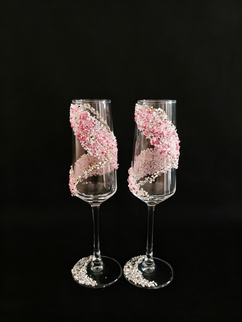 K&ouml;nnte beinhalten: Zwei Sektgl&auml;ser mit klarem Glas und einem dekorativen Design. Das Design zeigt eine Spirale aus rosa und wei&szlig;en Kristallen mit zus&auml;tzlichen Kristallen am Fu&szlig; jedes Glases. Der Hintergrund ist schwarz.