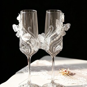 May include: Two clear champagne flutes, adorned with white butterfly and floral embellishments, silver rhinestones, and faux pearls. The stems are slender, and the bases are decorated with matching rhinestones and pearls. A decorative hairpiece is in the background.