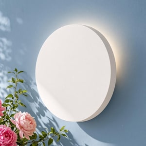 May include: A circular wall-mounted light fixture with a smooth, matte white finish. The lamp emits a soft, warm glow from behind its edges, creating a halo effect against the wall. The design is simple, flat, and geometric in its round shape.