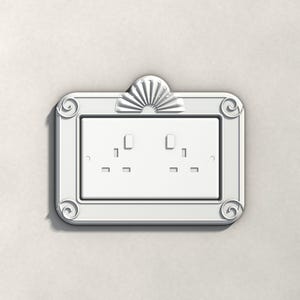 May include: A white double electrical wall socket faceplate featuring a rectangular frame with decorative scrollwork at each corner and a fan-shaped crest at the top centre. The plate is designed for a standard two-gang electrical socket.