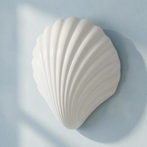 Peut inclure: Une pièce de décoration murale en céramique blanche en forme de coquille Saint-Jacques. L'objet présente une finition brillante avec des crêtes verticales profondes et rayonnantes qui s'étendent de la base étroite au bord supérieur large.