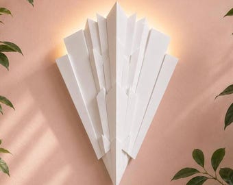 May include: A white wall sconce featuring a geometric, fan-like arrangement of layered, angular panels. The fixture emits a soft, warm glow from behind its structure, creating a sharp, pointed silhouette against the wall surface.