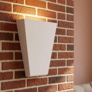 May include: A white, trapezoidal wall sconce mounted on a red brick surface. The fixture features a smooth, matte finish and emits a warm, upward-facing light that illuminates the textured brickwork directly above the lamp housing.
