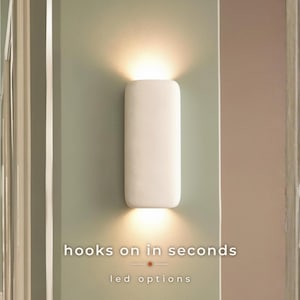 Peut inclure: Une applique murale rectangulaire blanche aux coins arrondis, diffusant une lumière chaude vers le haut et vers le bas. Le texte "hooks on in seconds" et "led options" est imprimé en blanc sous le luminaire sur la surface du mur.