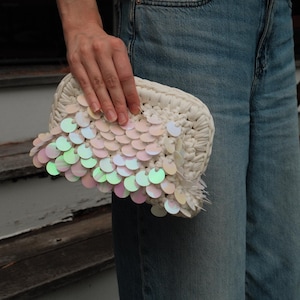 Puede incluir: Un pequeño bolso de mano de ganchillo en color blanco roto con una capa decorativa de lentejuelas circulares iridiscentes en tonos rosa pálido, verde claro y azul suave. El bolso tiene una forma redondeada y estructurada con textura.