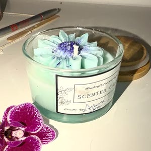 K&ouml;nnte beinhalten: Duftkerze in einem transparenten Glas, mit hellblauem Blumendesign. Die Kerze hat einen hellblauen Farbton und ein Etikett mit der Aufschrift "Scented Candle". Eine lila Orchidee befindet sich im Vordergrund.