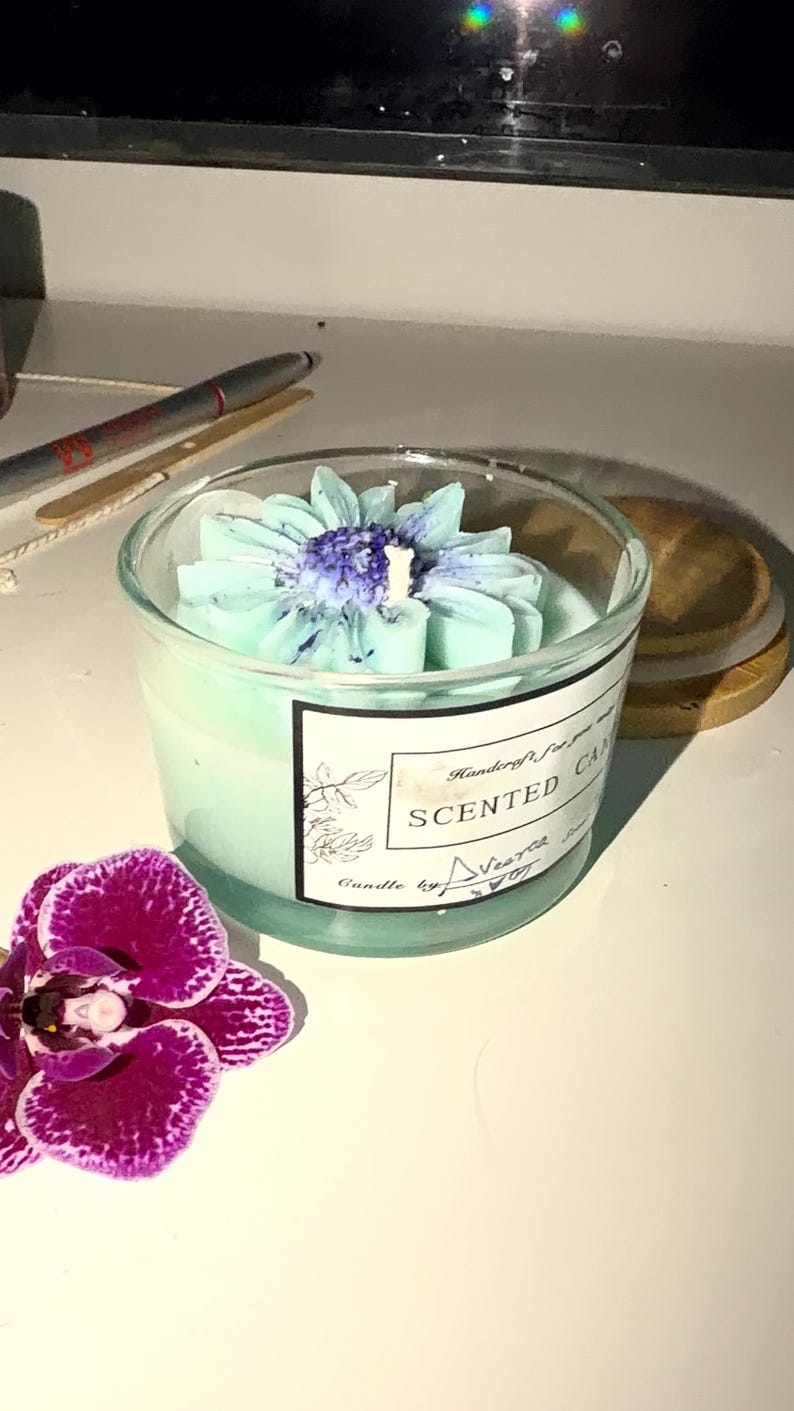 K&ouml;nnte beinhalten: Duftkerze in einem transparenten Glas, mit hellblauem Blumendesign. Die Kerze hat einen hellblauen Farbton und ein Etikett mit der Aufschrift "Scented Candle". Eine lila Orchidee befindet sich im Vordergrund.
