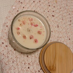 K&ouml;nnte beinhalten: Ein klares Glas-Kerzenglas, gef&uuml;llt mit wei&szlig;em Wachs und mit kleinen rosa Bl&uuml;ten dekoriert. Ein Holzdeckel liegt neben dem Glas auf einer rosa Paillettenoberfl&auml;che. Die Kerze ist ein dekorativer Artikel f&uuml;r die Wohnung.