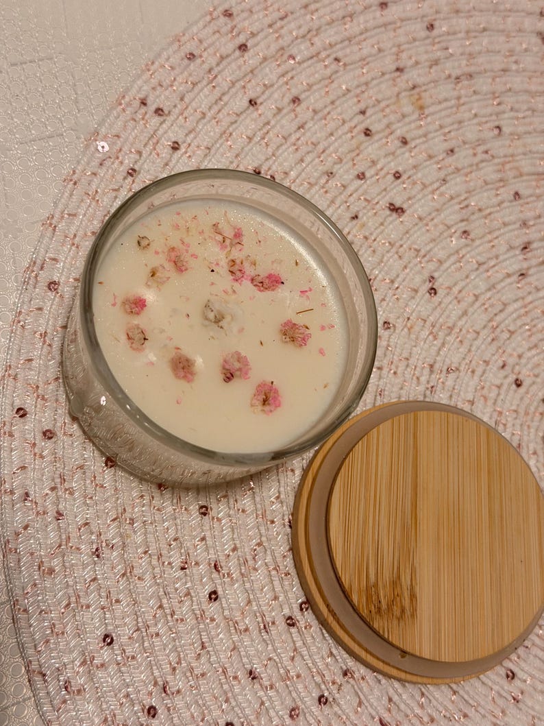 K&ouml;nnte beinhalten: Ein klares Glas-Kerzenglas, gef&uuml;llt mit wei&szlig;em Wachs und mit kleinen rosa Bl&uuml;ten dekoriert. Ein Holzdeckel liegt neben dem Glas auf einer rosa Paillettenoberfl&auml;che. Die Kerze ist ein dekorativer Artikel f&uuml;r die Wohnung.