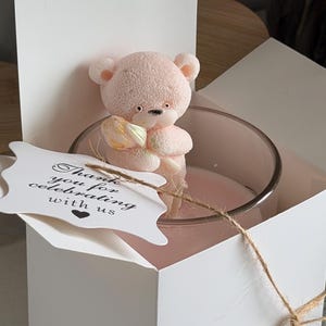 K&ouml;nnte beinhalten: Eine rosa Teddyb&auml;r-Kerze in einem transparenten Glasbeh&auml;lter, pr&auml;sentiert in einer wei&szlig;en Geschenkbox. Ein Anh&auml;nger mit dem Text "Thank you for celebrating with us" ist mit Bindfaden befestigt. Die Kerze ist hellrosa.
