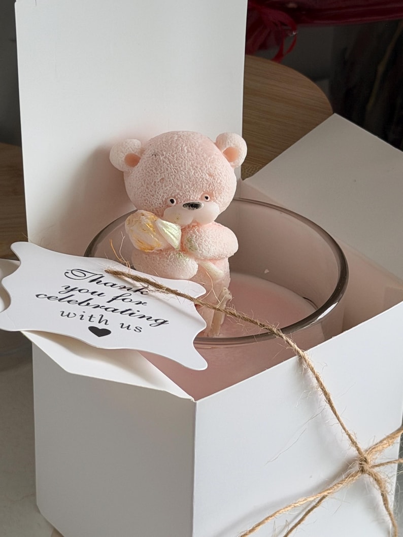 K&ouml;nnte beinhalten: Eine rosa Teddyb&auml;r-Kerze in einem transparenten Glasbeh&auml;lter, pr&auml;sentiert in einer wei&szlig;en Geschenkbox. Ein Anh&auml;nger mit dem Text "Thank you for celebrating with us" ist mit Bindfaden befestigt. Die Kerze ist hellrosa.