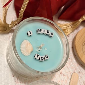 K&ouml;nnte beinhalten: Ein klarer Glashalter mit einer hellblauen Kerze. Die Kerze tr&auml;gt die Worte "MISS YOU", die mit kleinen Buchstabenperlen geschrieben sind. Eine Muschel und dekorative Elemente sind in die Kerze eingebettet.