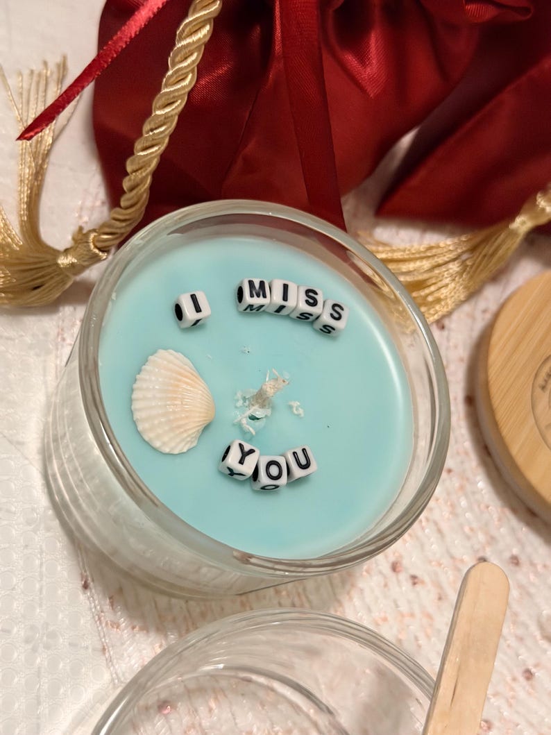 K&ouml;nnte beinhalten: Ein klarer Glashalter mit einer hellblauen Kerze. Die Kerze tr&auml;gt die Worte "MISS YOU", die mit kleinen Buchstabenperlen geschrieben sind. Eine Muschel und dekorative Elemente sind in die Kerze eingebettet.