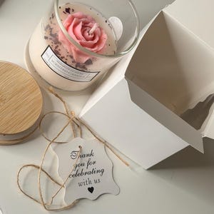 K&ouml;nnte beinhalten: Eine Duftkerze in einem transparenten Glas mit einem rosafarbenen, rosenf&ouml;rmigen Wachsdesign. Die Kerze wird von einem Holzdeckel, einem Geschenkanh&auml;nger mit der Aufschrift "Thank you for celebrating with us" und einer wei&szlig;en Box begleitet.