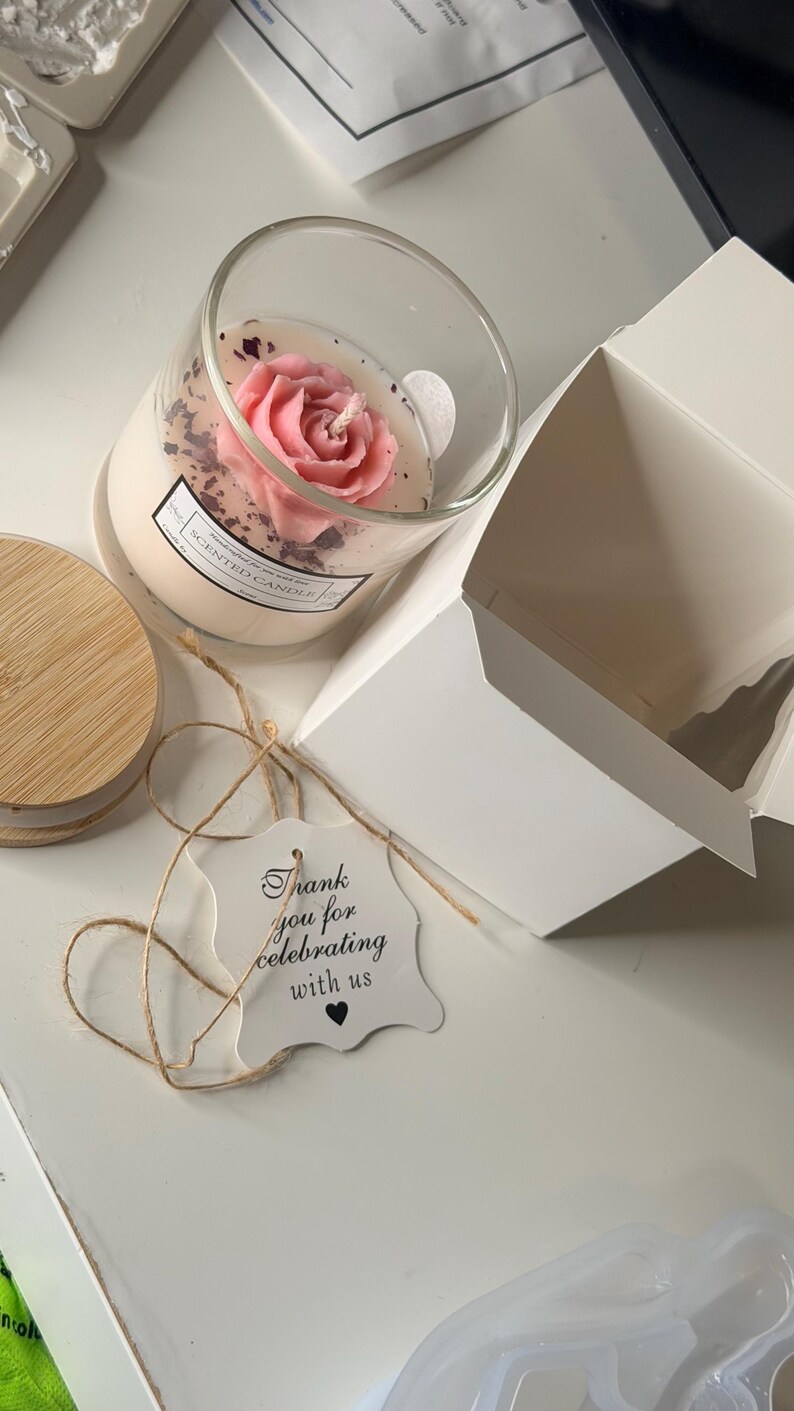 K&ouml;nnte beinhalten: Eine Duftkerze in einem transparenten Glas mit einem rosafarbenen, rosenf&ouml;rmigen Wachsdesign. Die Kerze wird von einem Holzdeckel, einem Geschenkanh&auml;nger mit der Aufschrift "Thank you for celebrating with us" und einer wei&szlig;en Box begleitet.
