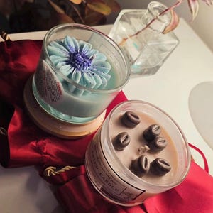 K&ouml;nnte beinhalten: Zwei Duftkerzen in Gl&auml;sern. Eine Kerze ist hellblau und blumenf&ouml;rmig. Die andere ist beige mit Kaffeebohnen-Akzenten. Beide Kerzen stehen auf einer roten Oberfl&auml;che.