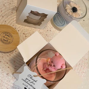 K&ouml;nnte beinhalten: Duftkerzen in Gl&auml;sern, eine mit einem rosa Teddyb&auml;ren und eine mit einer Blume. Die Kerzen sind in wei&szlig;en Schachteln mit einem Fenster und einem Anh&auml;nger mit der Aufschrift "Thank you for celebrating with us" verpackt.