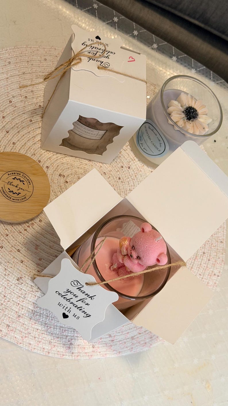 K&ouml;nnte beinhalten: Duftkerzen in Gl&auml;sern, eine mit einem rosa Teddyb&auml;ren und eine mit einer Blume. Die Kerzen sind in wei&szlig;en Schachteln mit einem Fenster und einem Anh&auml;nger mit der Aufschrift "Thank you for celebrating with us" verpackt.