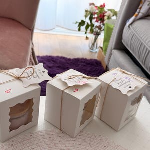 K&ouml;nnte beinhalten: Drei wei&szlig;e quadratische Geschenkboxen mit transparenten Fensterausschnitten, mit Bindfaden gebunden und mit dekorativen Anh&auml;ngern verziert. Jede Box hat eine Nummer. Die Anh&auml;nger tragen den Text "Vielen Dank, dass Sie mit uns feiern."