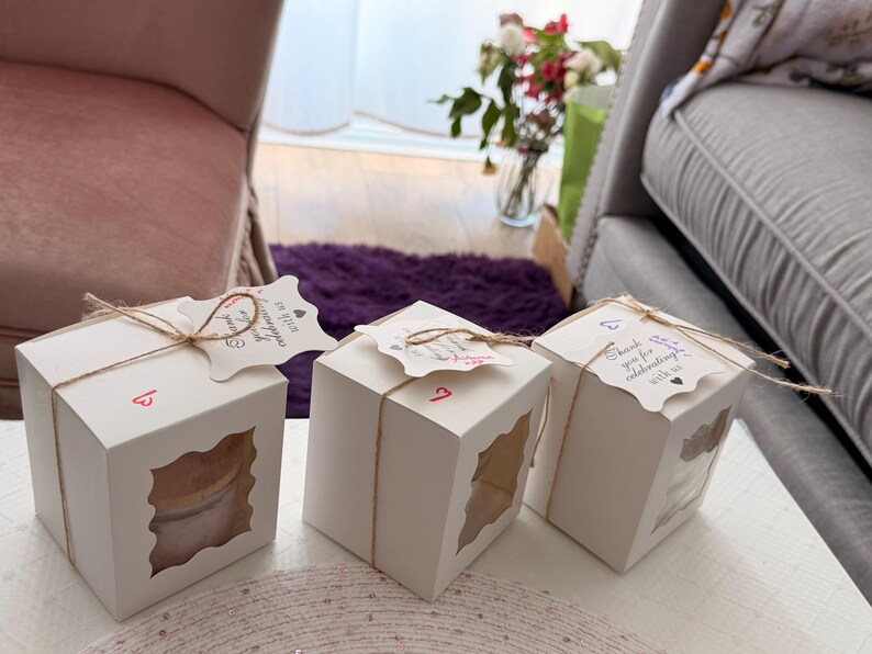 K&ouml;nnte beinhalten: Drei wei&szlig;e quadratische Geschenkboxen mit transparenten Fensterausschnitten, mit Bindfaden gebunden und mit dekorativen Anh&auml;ngern verziert. Jede Box hat eine Nummer. Die Anh&auml;nger tragen den Text "Vielen Dank, dass Sie mit uns feiern."