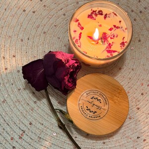 K&ouml;nnte beinhalten: Eine brennende Kerze in einem durchsichtigen Glas mit getrockneten Rosenbl&auml;ttern. Ein Holzdeckel mit dem Text "Thank you" und "Aveerah Candles" befindet sich neben der Kerze. Eine getrocknete burgunderfarbene Rose ist ebenfalls im Bild.