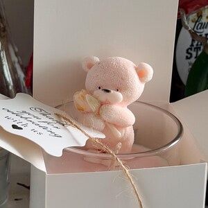 K&ouml;nnte beinhalten: Eine rosa Teddyb&auml;r-Kerze, die eine Blume h&auml;lt, in einem klaren Glas, pr&auml;sentiert in einer wei&szlig;en Geschenkbox. Ein Anh&auml;nger mit dem Text "Thank you for celebrating with us" ist mit Bindfaden befestigt. Die Kerze ist ein dekorativer Artikel.