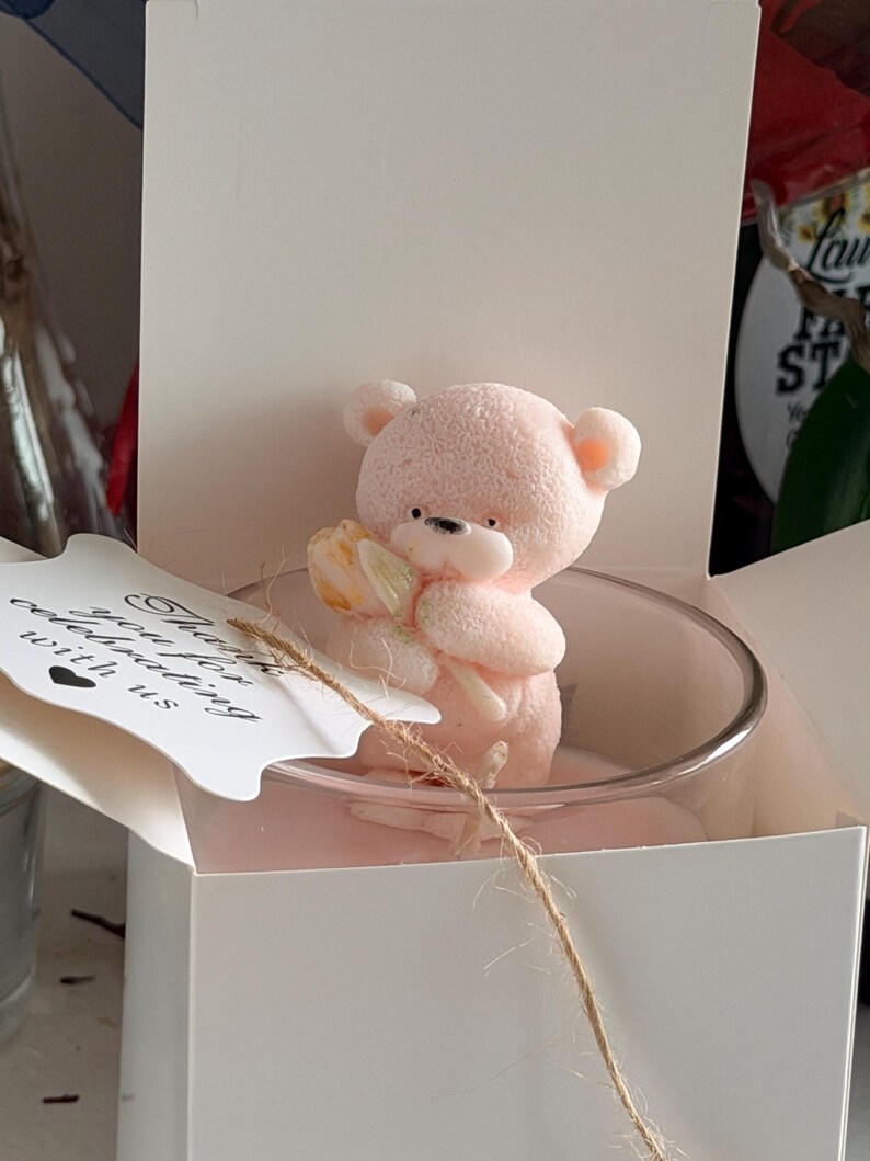 K&ouml;nnte beinhalten: Eine rosa Teddyb&auml;r-Kerze, die eine Blume h&auml;lt, in einem klaren Glas, pr&auml;sentiert in einer wei&szlig;en Geschenkbox. Ein Anh&auml;nger mit dem Text "Thank you for celebrating with us" ist mit Bindfaden befestigt. Die Kerze ist ein dekorativer Artikel.