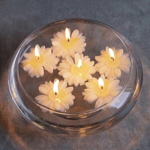 K&ouml;nnte beinhalten: Eine klare Glasschale mit Wasser, in der sechs wei&szlig;e, schwimmende Kerzen in Form von G&auml;nsebl&uuml;mchen schwimmen. Jede Kerze hat eine gelbe Mitte und einen brennenden Docht, der ein warmes Leuchten erzeugt. Die Schale steht auf einer grauen Oberfl&auml;che.