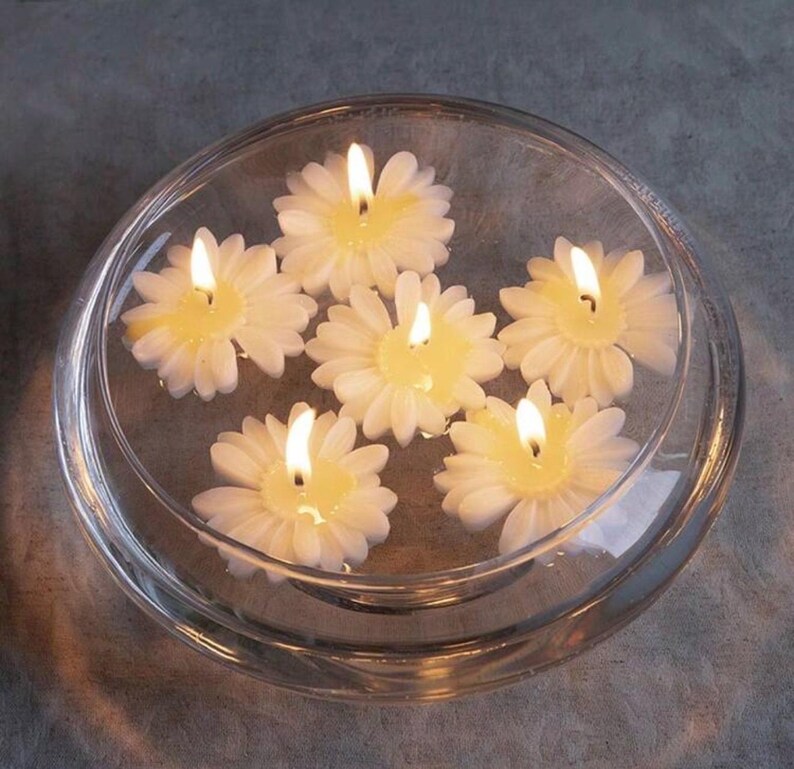 K&ouml;nnte beinhalten: Eine klare Glasschale mit Wasser, in der sechs wei&szlig;e, schwimmende Kerzen in Form von G&auml;nsebl&uuml;mchen schwimmen. Jede Kerze hat eine gelbe Mitte und einen brennenden Docht, der ein warmes Leuchten erzeugt. Die Schale steht auf einer grauen Oberfl&auml;che.