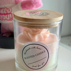 K&ouml;nnte beinhalten: Duftkerze in einem transparenten Glas, gekr&ouml;nt von einem hellrosa Blumendesign. Die Kerze hat einen Holzdeckel und ein Etikett mit der Aufschrift "Handmade with love, Scented Candle, just for you."