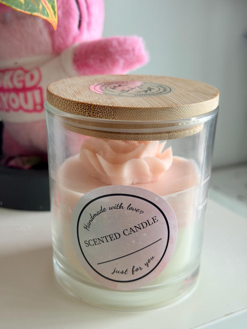 K&ouml;nnte beinhalten: Duftkerze in einem transparenten Glas, gekr&ouml;nt von einem hellrosa Blumendesign. Die Kerze hat einen Holzdeckel und ein Etikett mit der Aufschrift "Handmade with love, Scented Candle, just for you."