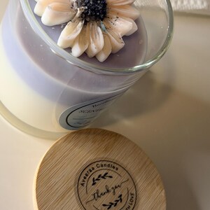 K&ouml;nnte beinhalten: Eine dekorative Kerze in einem transparenten Glas, gekr&ouml;nt von einer dekorativen Blume. Die Kerze hat ein hellviolettes und wei&szlig;es Farbschema. Ein Holzdeckel mit dem Text "Aveeraa Candles" und "Thank you" liegt neben der Kerze.