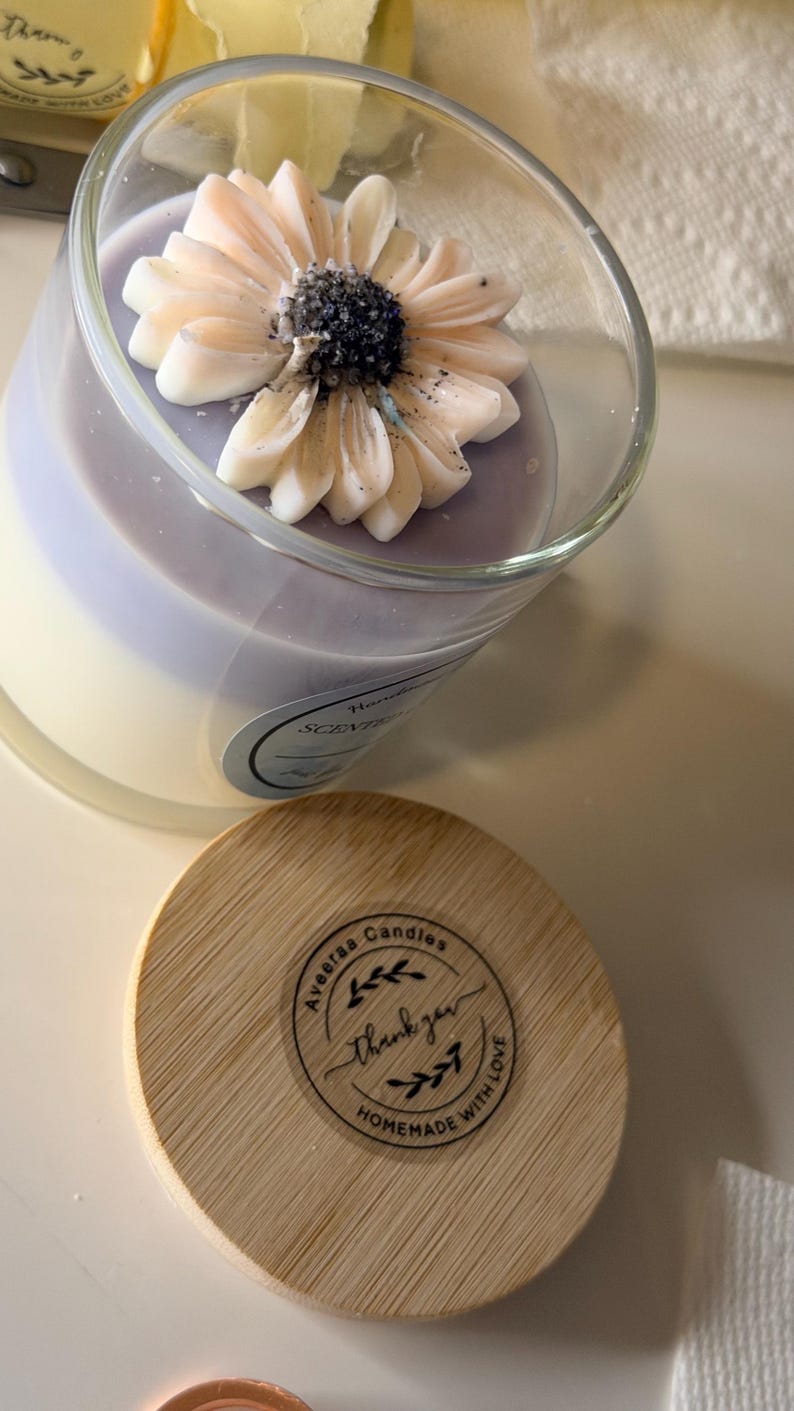 K&ouml;nnte beinhalten: Eine dekorative Kerze in einem transparenten Glas, gekr&ouml;nt von einer dekorativen Blume. Die Kerze hat ein hellviolettes und wei&szlig;es Farbschema. Ein Holzdeckel mit dem Text "Aveeraa Candles" und "Thank you" liegt neben der Kerze.